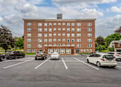 Thumbnail 1 of 7 - A parking lot in front of a red brick building with cars parked.