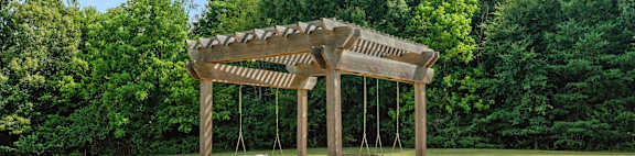 A wooden pergola with two benches is situated in a grassy area.