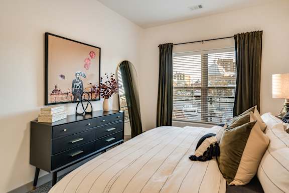 A bedroom with a large bed, a painting on the wall, and a window with blinds.at Converge KC Uptown, Missouri