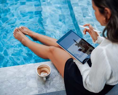 A woman is sitting by a pool with a tablet in her lap and a cup of coffee in front of her.