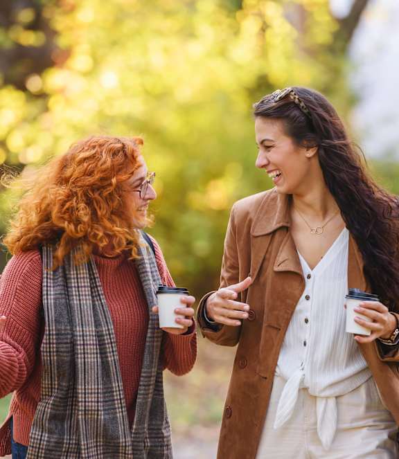 Two women are walking and talking while holding coffee cups.