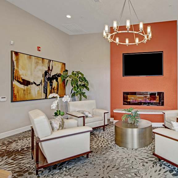 The amazing resident lounge with a fireplace, plush chairs, decorations, TV mounted on an orange accent wall, plush chairs, and a coffee table at Vista View Apartment Homes. 