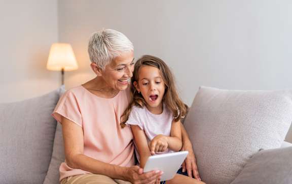 An older woman and a younger girl are sitting on a couch looking at a tablet.