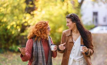 Two women are walking and talking while holding coffee cups.