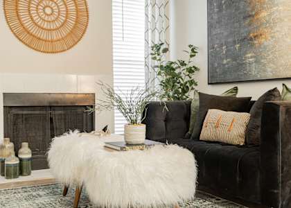 A living room with a fireplace, a white fluffy ottoman, a black couch, and a large painting on the wall.