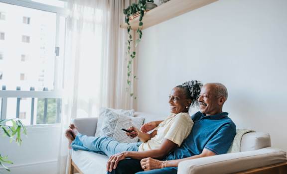 A couple is sitting on a couch in a bright room with a window and a plant.
