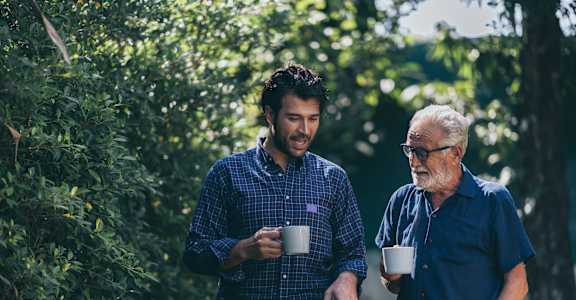 Two men are standing in a green area, one holding a cup and the other holding a mug.