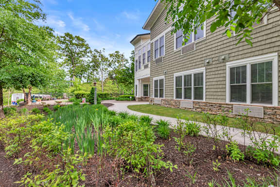 A building with a grey siding and white trim is surrounded by a garden with green plants.