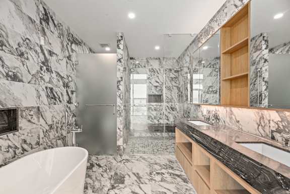 A bathroom with a marble patterned floor and walls, a white bathtub, and a wooden shelf. at Olympic + Hill Apartments, Apartments, Los Angeles, CA