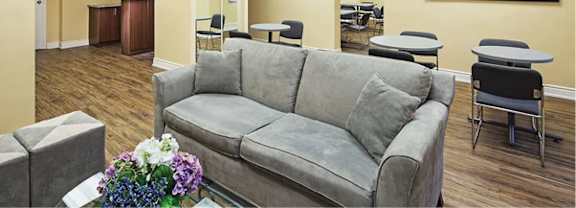 A grey couch with a pillow is in the middle of a room with a coffee table and a vase of flowers.