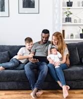 A family of three is sitting on a grey couch with a tablet in the man's hands.
