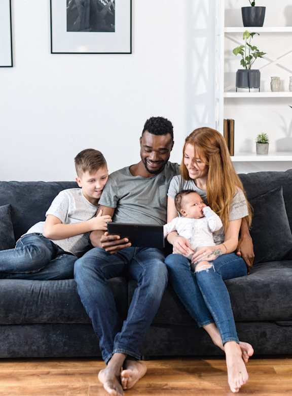 A family of four is sitting on a grey couch with a baby in the woman's arms.