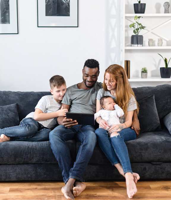 A family of four is sitting on a grey couch with a baby in the woman's arms.