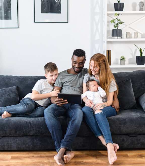A family of three is sitting on a grey couch with a tablet in the man's hands.