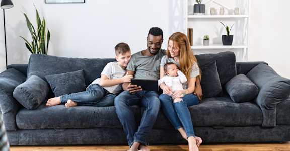 A family of three is sitting on a grey couch with a baby in the woman's arms.