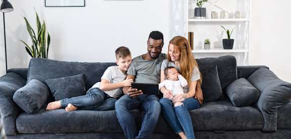 A family of four is sitting on a grey couch with a baby in the woman's arms.