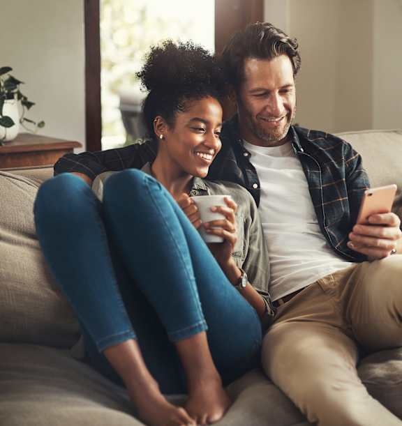 A man and a woman are sitting on a couch, both looking at a smartphone.