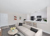 Thumbnail 5 of 9 - A living room with a couch, coffee table, and kitchen area in the background.