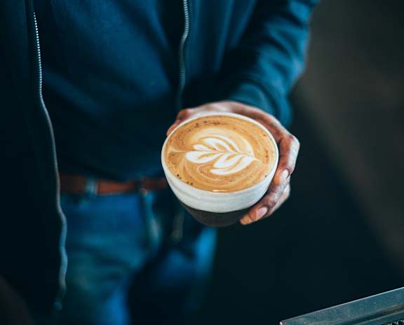 A person in a blue shirt is holding a cup of coffee with a latte art design on it.