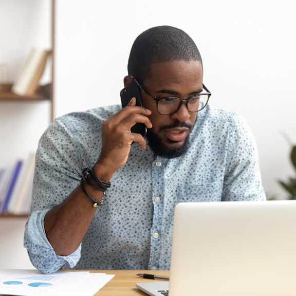 A man in a blue shirt is talking on the phone while sitting at a desk with a laptop and some papers.