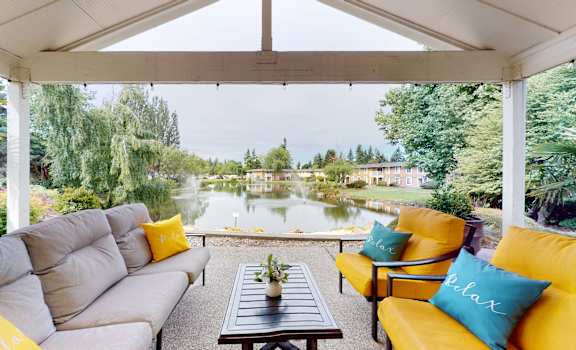 A patio with a gray couch and orange chairs with green trees and a pond in the background at Lakeside Apartments. 