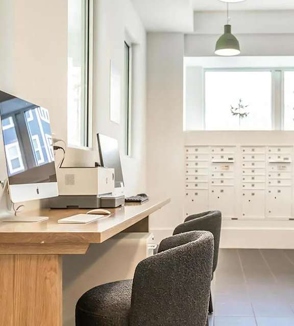 Business center with two desktop computers, a printer and upholstered chairs at a long desk, beside a wall of mailboxes.