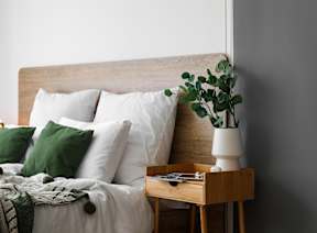 A bed with white pillows and a green throw is next to a wooden nightstand with a plant on it.