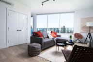 A living room with a grey couch, a wicker ottoman, and a white rug.