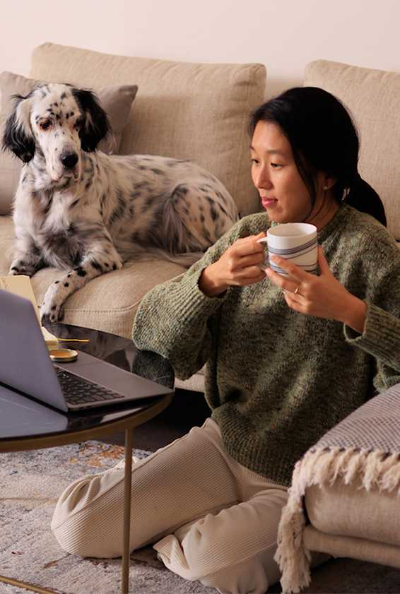 A woman in a green sweater is sitting on the floor with a laptop and a cup in her hand, while a dog sits on the couch behind her.