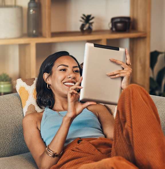 A woman is sitting on a grey couch holding a tablet.
