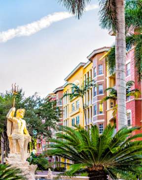 A statue of a man holding a woman is in the middle of a park with palm trees and buildings in the background.