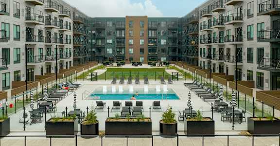 A large pool surrounded by lounge chairs and planters is surrounded by apartment buildings.
