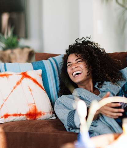 A woman with curly hair is laughing while sitting on a couch.