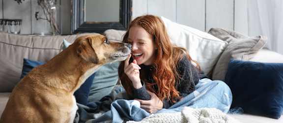 A woman is sitting on a bed with a dog licking her face.