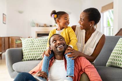 A family of three is sitting on a couch, with the child on the man's shoulders.
