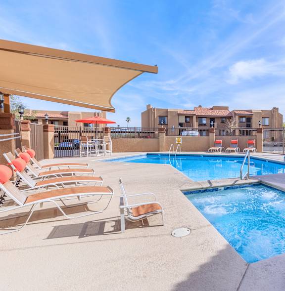 A pool with chairs and a sunshade is surrounded by buildings.
