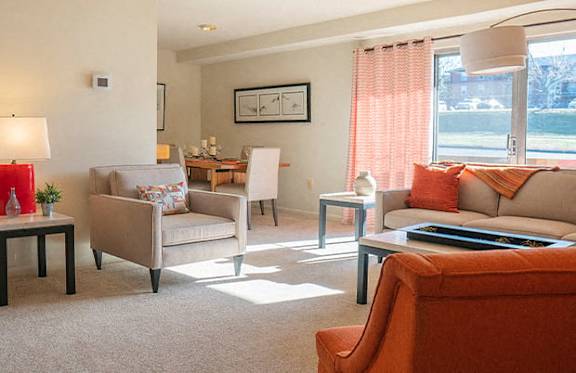 A living room with a grey couch, a red couch, a white chair, a small table with a vase and a plant on it, and a window with orange curtains.