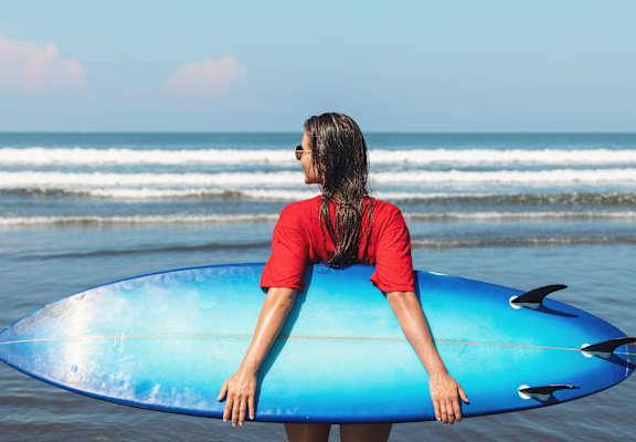 A person in a red shirt is holding a blue surfboard at the beach.