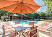 Thumbnail 3 of 27 - Swimming pool with striped lounge chairs under orange umbrella at The Colony Apartments, Texas, 78148