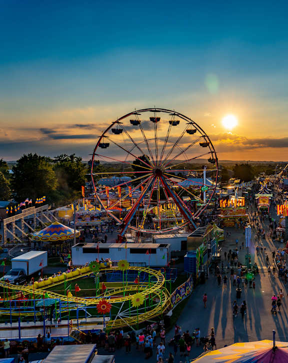 A sunset view of a bustling fair with a Ferris wheel and other rides.