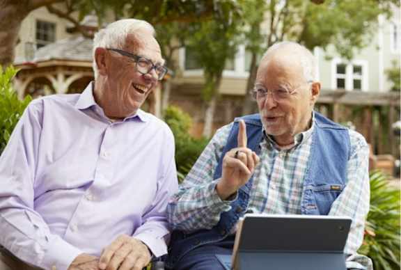 Two older men are sitting outside, one is holding a laptop.