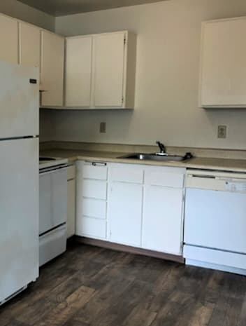 A white refrigerator stands in a kitchen with white cabinets and a wooden floor.