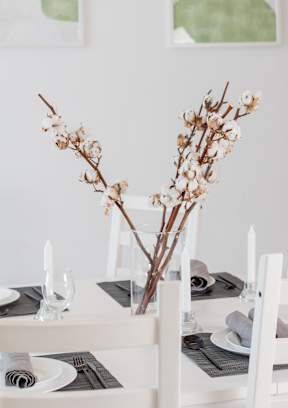 A table set for a meal with white chairs and a vase of dried flowers in the center.