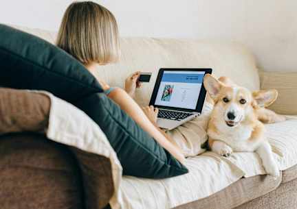 A woman is sitting on a couch with a laptop and a tablet, accompanied by a corgi dog.