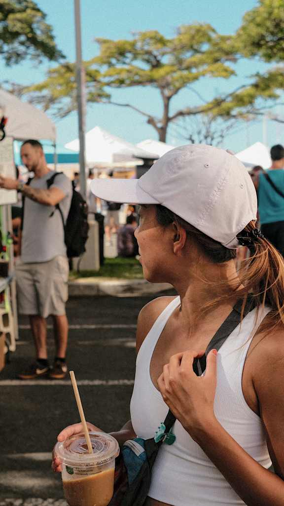 A woman in a white tank top and white cap is holding a cup with a straw in it.