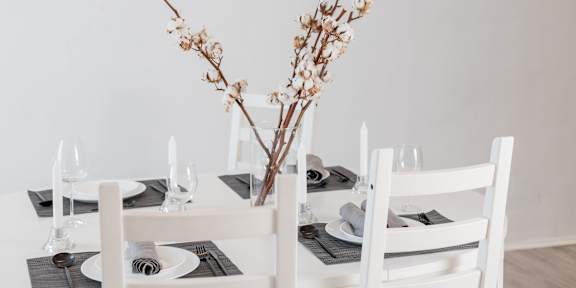 A table set for a meal with white chairs and a vase of dried flowers in the center.