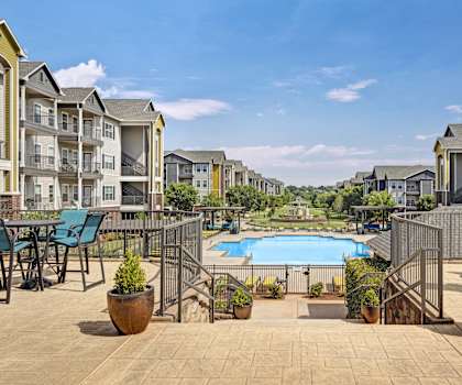 The turquoise outdoor pool with pool chairs, tables, and chairs with the apartments in the background at Lincoln at Central Park Apartment Homes. 