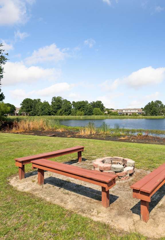 Two red benches are in the foreground of a grassy area with a body of water and buildings in the background.