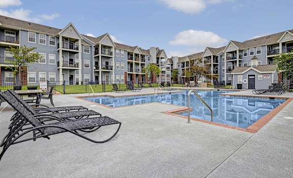 The turquoise outdoor pool with pool chairs, tables, and chairs with the apartments in the background at Harbin Pointe Apartment Homes. 