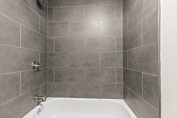 A white bathtub sits in a bathroom with grey tiled walls.